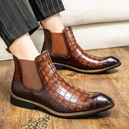 A person wearing MVPs' Chelsea Leather Boots, crafted from high-quality brown split leather with a crocodile pattern, featuring black soles and elastic side panels, pairs them elegantly with pinstriped trousers while standing on a wooden floor.